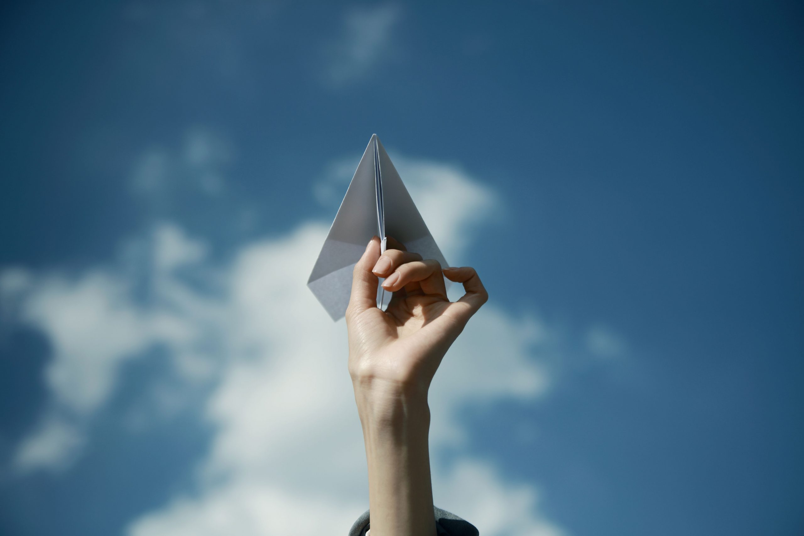Hand holding a paper airplane with blue skies and clouds in the background